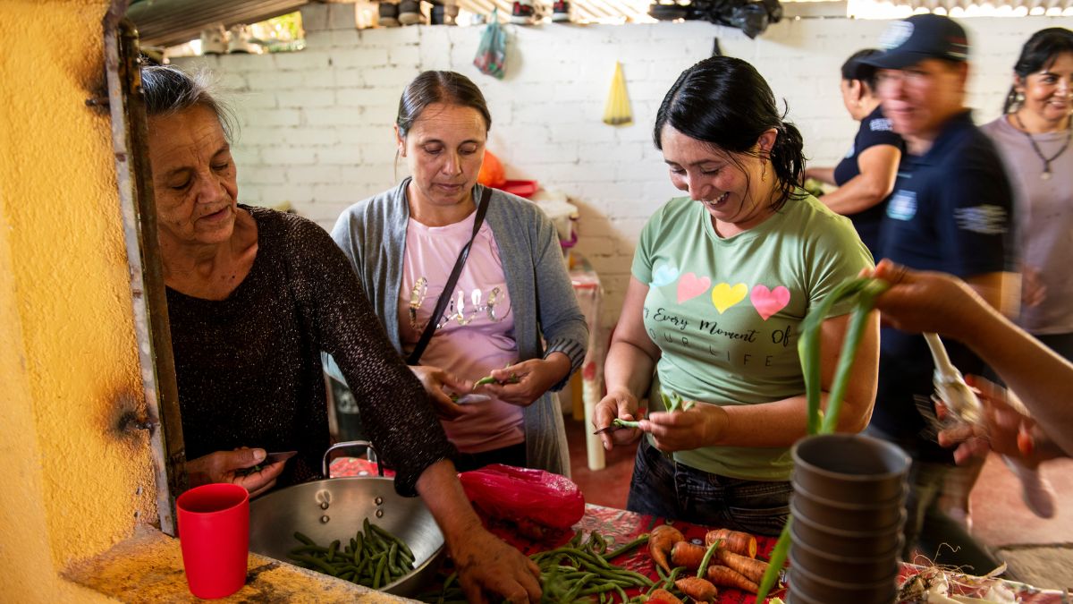 Drei Frau schneiden gemeinsam Gemüse, dahinter stehen drei weitere Leute