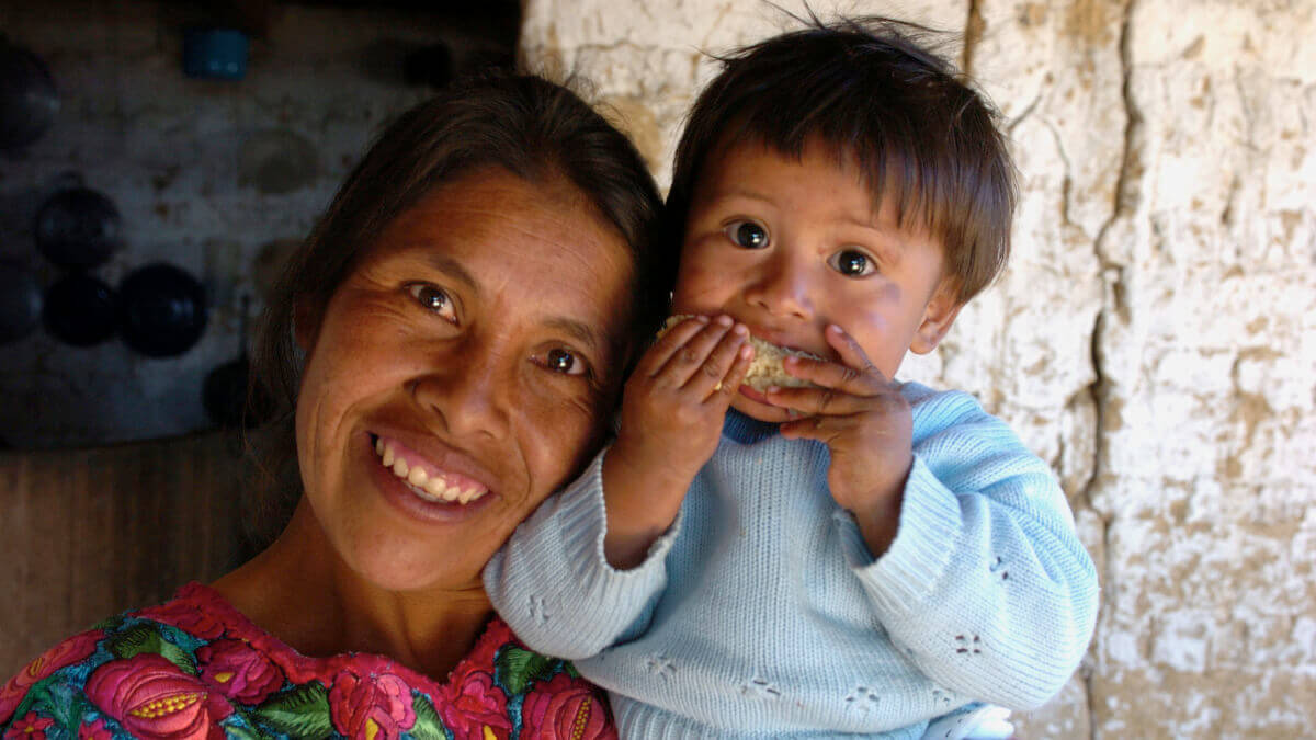 Eine lachende Frau hält ein kleines Kind auf dem Arm, welches einen Maiskolben ißt.